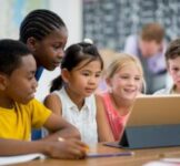 A multi-ethnic group of elementary age children are sitting together and are watching an educational video on a digital tablet.