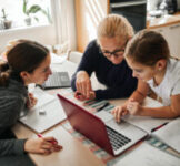 Mother helping her daughters to finish school homework during coronavirus quarantine. They are using laptop.